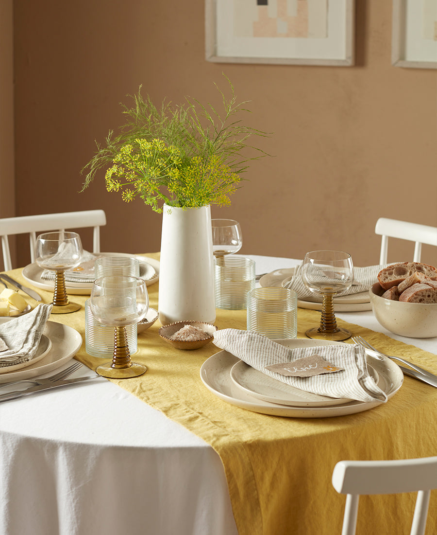 A round table is laid with white table cloth yellow linen runner and yellow flowers.